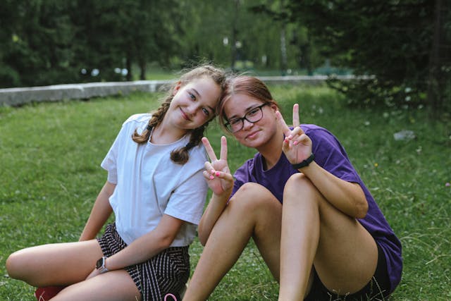 Two girls sitting on the grass outdoors and making peace signs at the camera 