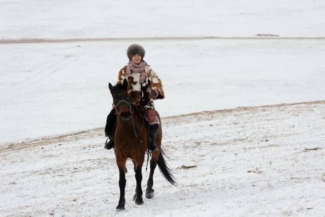 Mongolian rider wearing traditional winter clothing while travelling on horseback through the snow.