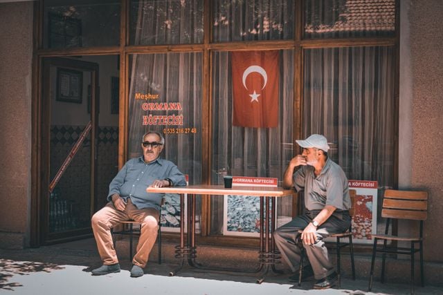 Two men sitting outside a small Turkish restaurant, offering a scene of everyday spoken Turkish in the local community.