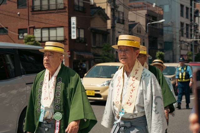 Two elderly Japanese men wearing traditional clothing and straw hats walking in a street procession.