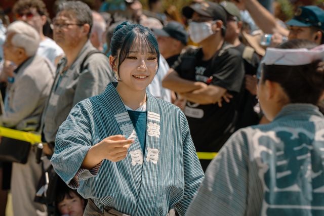 A smiling Japanese woman in traditional festival clothing speaking with another participant in a crowd.