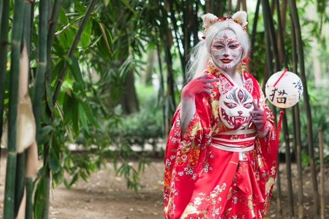 Person dressed as a Japanese fox spirit in a red kimono, wearing traditional makeup and holding a fox mask in a bamboo forest