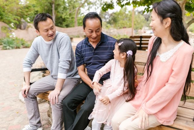 Three generations of a Korean family sitting together on a park bench, smiling and enjoying a conversation outdoors.